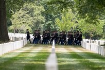 Army Soldiers dressed in dark ceremonial uniforms are seen marching in formation in the distance near rows of white tombstones.