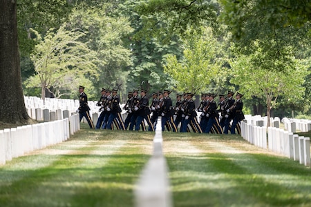 Army Soldiers dressed in dark ceremonial uniforms are seen marching in formation in the distance near rows of white tombstones.