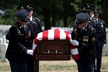 Army Soldiers dressed in dark ceremonial uniforms are carrying a brown, wooden, US flag-draped casket through Arlington National Cemetery.