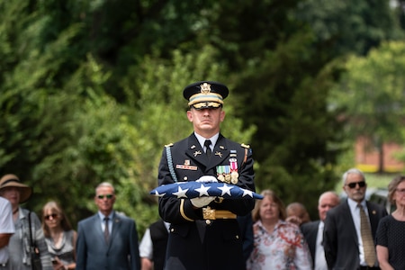 Army soldier in ceremonial dress uniform is holding a folded US flag in front of his check with both hands while others in civilian clothes are behind him watching a funeral service.