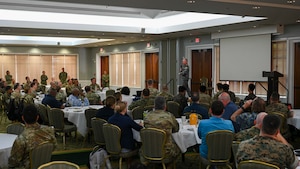An older man in military uniform stands on a stage facing a crowd sitting at round tables.