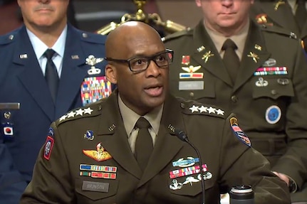 A man in a military uniform and eyeglasses sits behind a microphone delivering testimony.