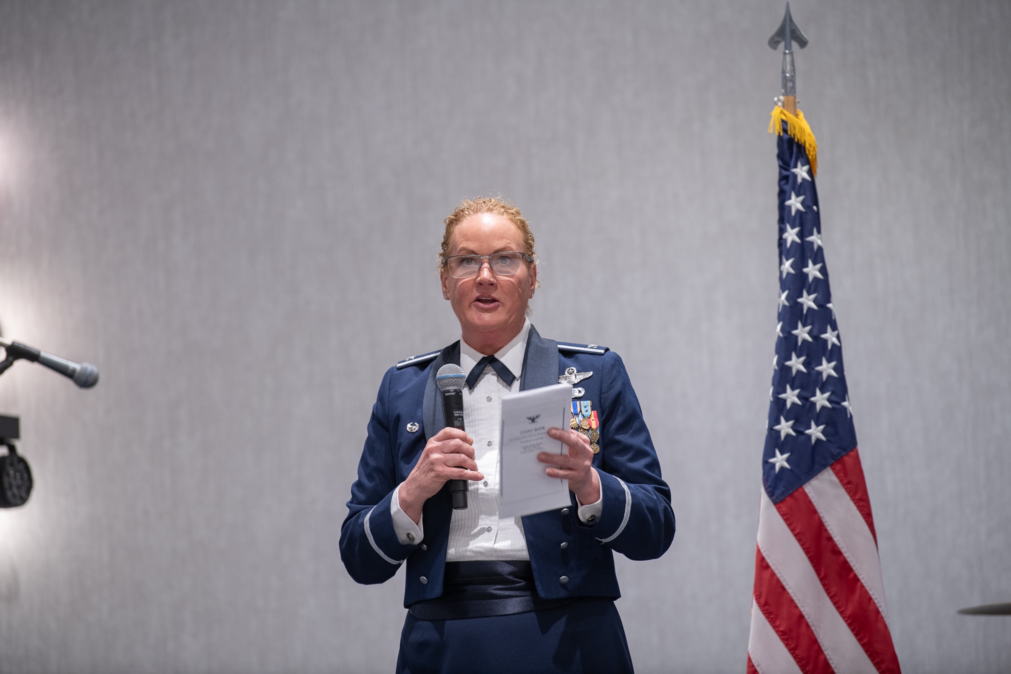 A servicemember stands next to an American flag while holding a microphone and piece of paper.