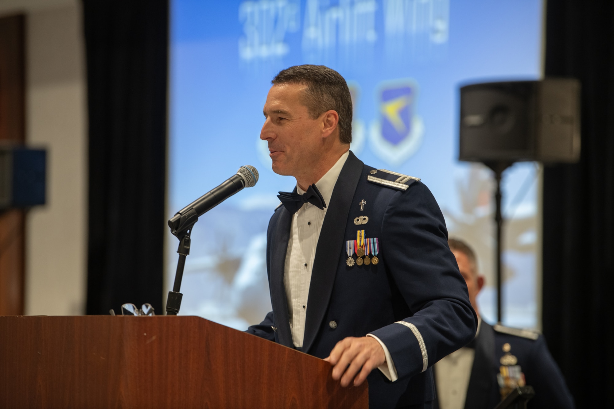 A servicemember stands at a podium in a ballroom with a slide presentation in the background.