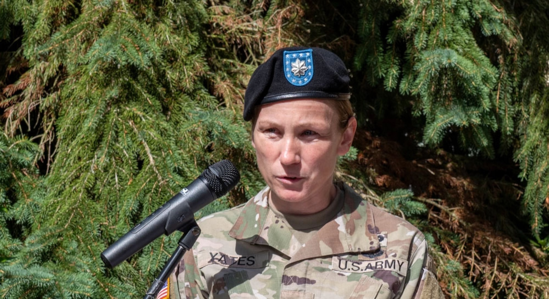 A woman wearing a black beret and a Army OCP uniform speaks at a podium outside.