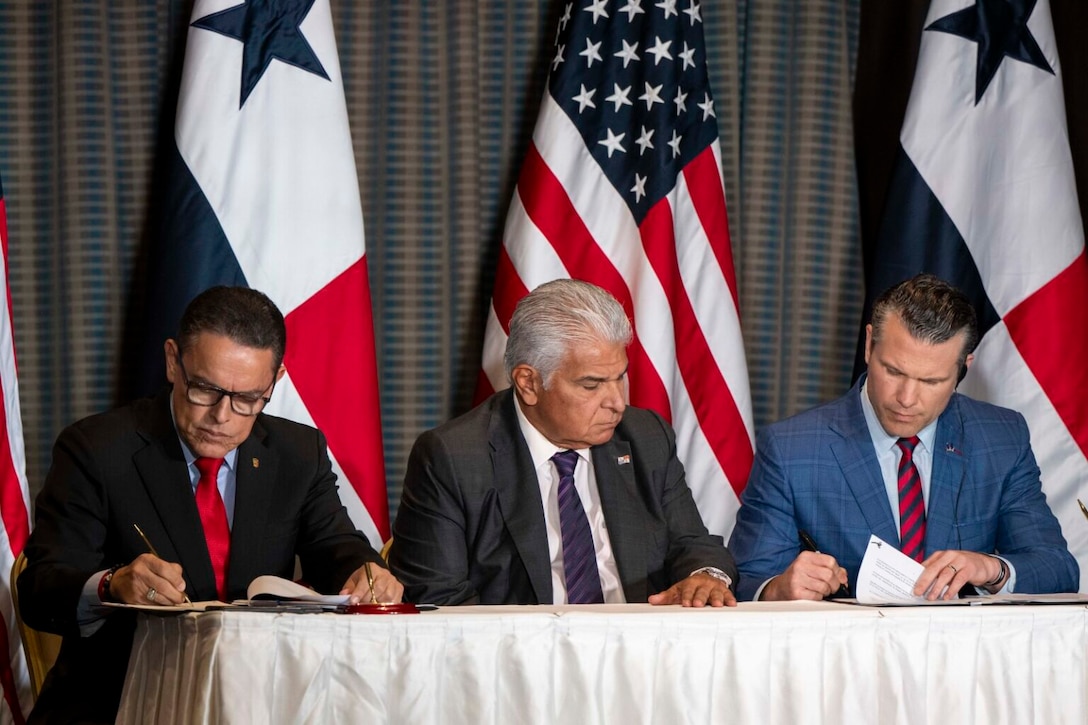 Three men sit at a table and sign documents.