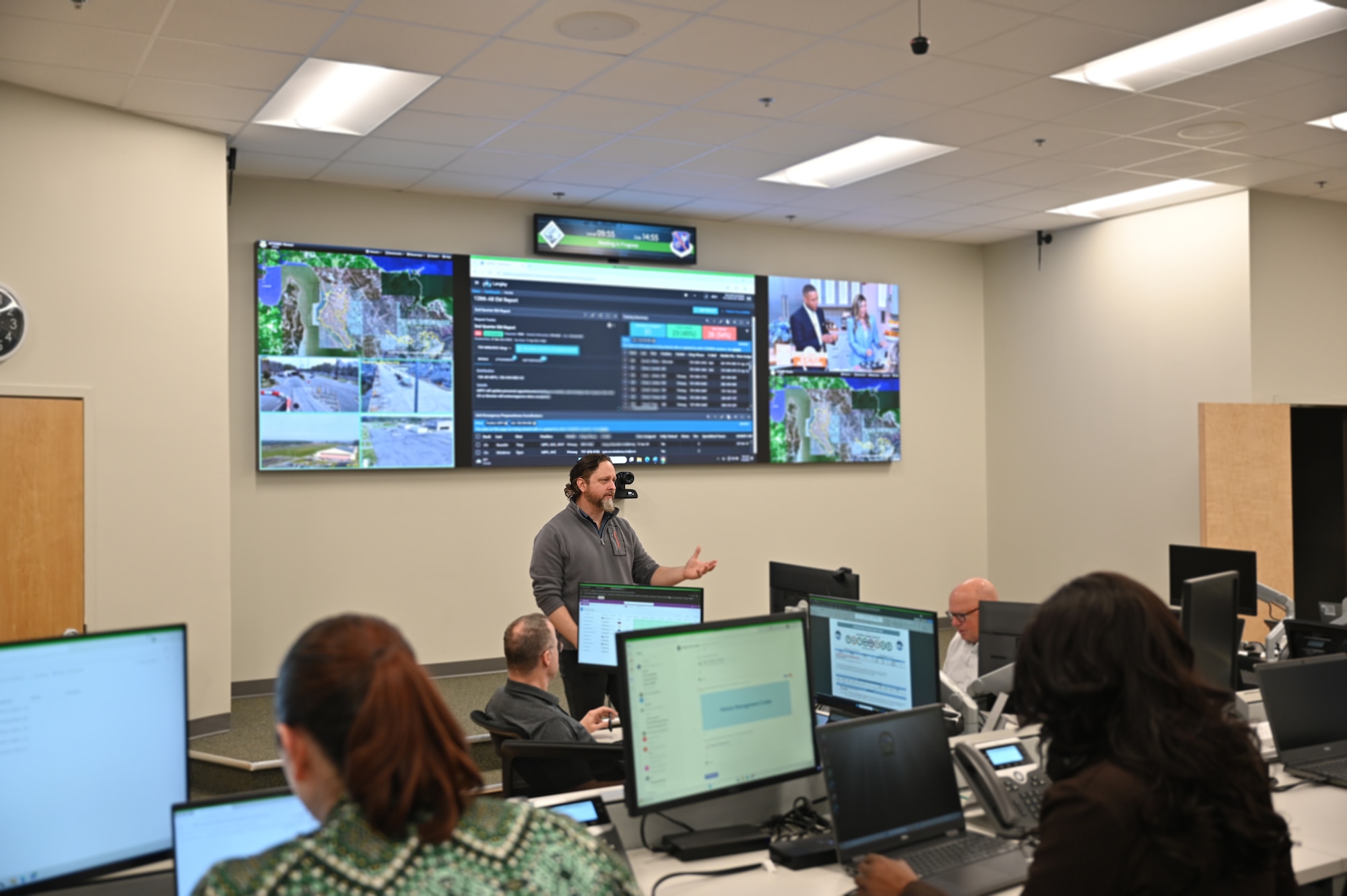 Man stands in front of a room of people surrounded by high tech screens. He is speaking to the people in the room.