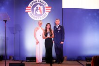 Three people stand on stage with the awardee standing in the center holding an award.