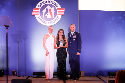 Three people stand on stage with the awardee standing in the center holding an award.