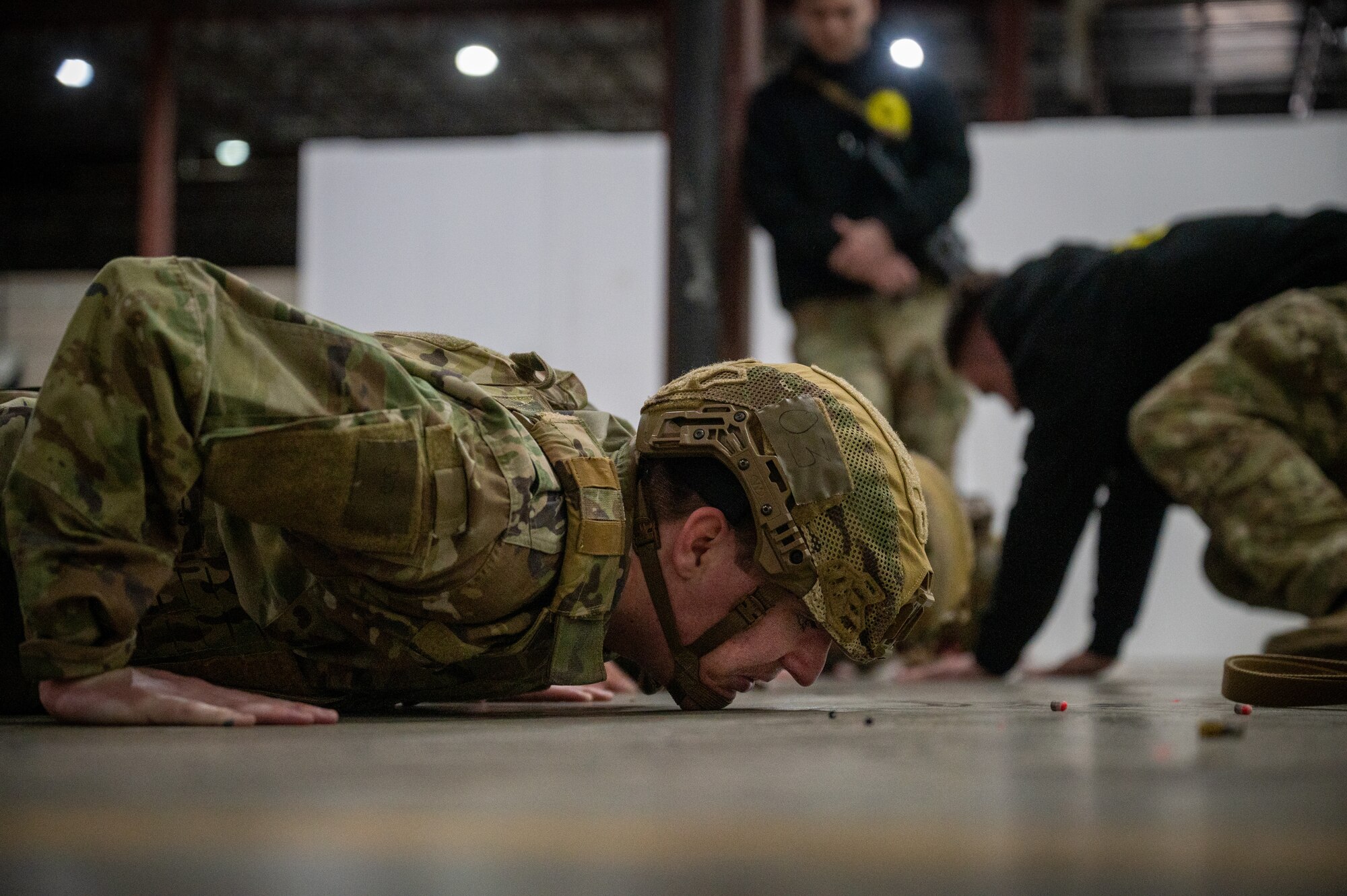 a line of people lay on the ground on their stomachs, with hands positioned to push themselves into a pushup, as two instructors look on