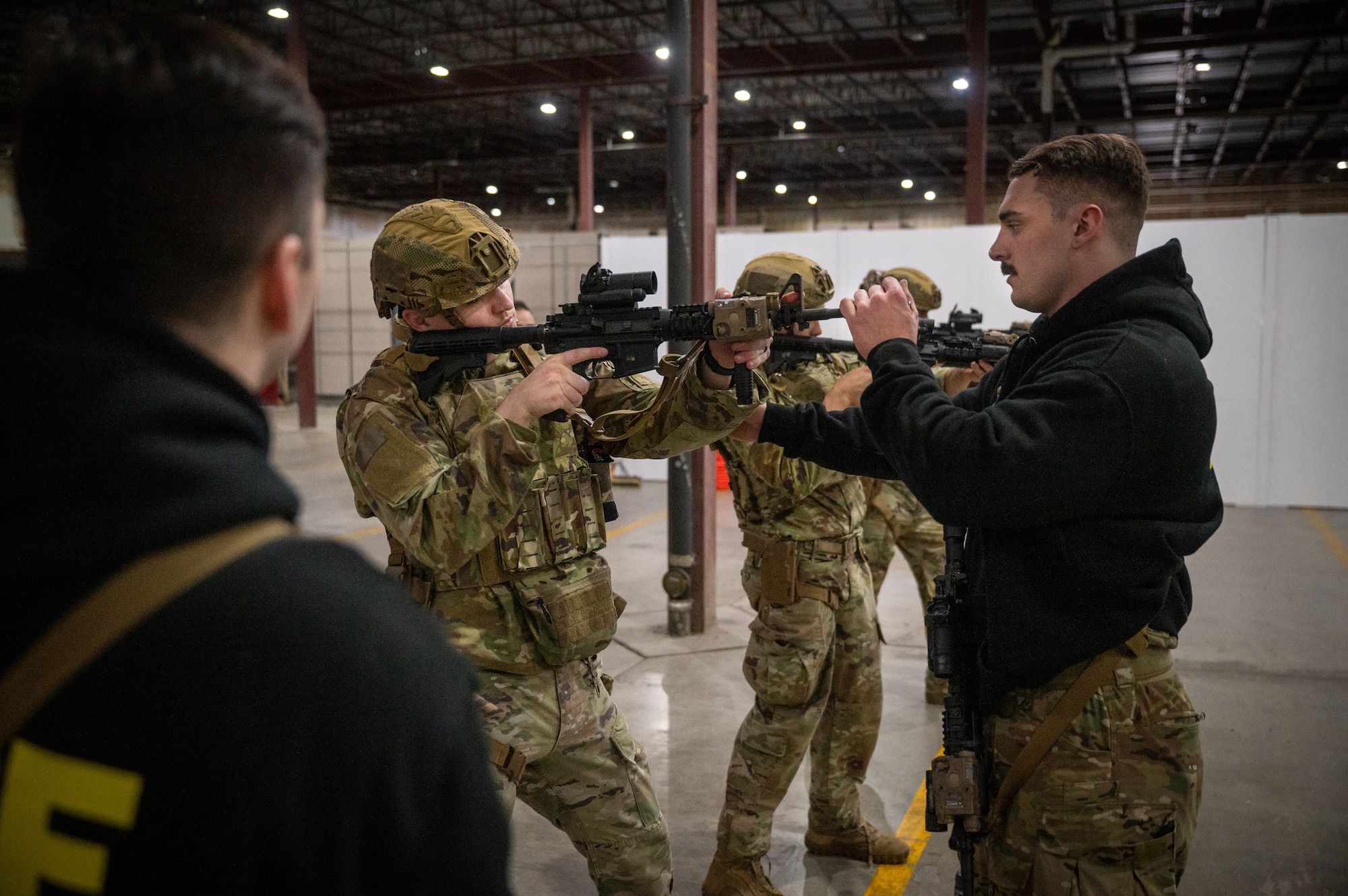 one man in fatigues and a helmet holds a fake weapon at the ready as another man adjusts his grip and aim