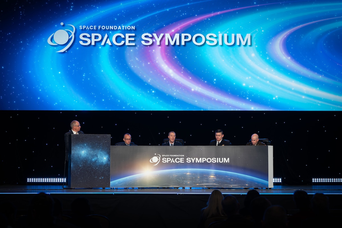 One moderator and 4 panelists are on an elevated stage. The moderator stands at a podium to the left, with the 4 panelists seated to the right.