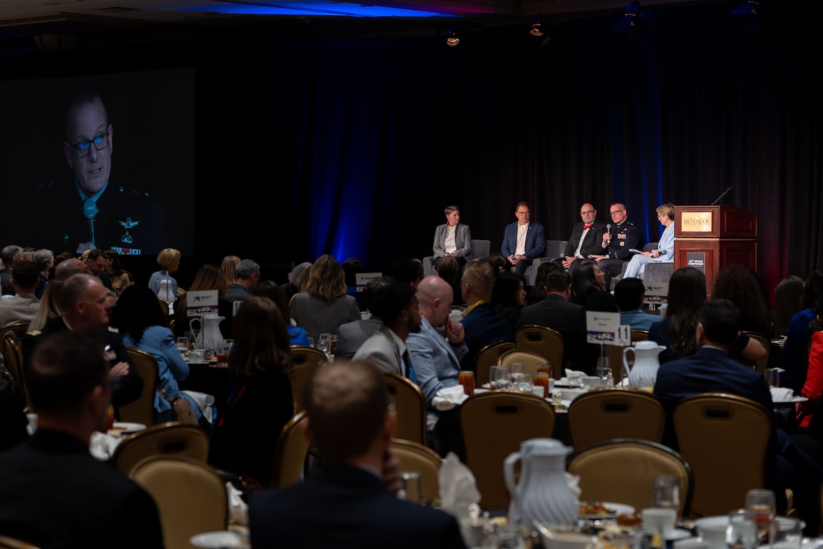 5 panelists sit on a stage with audience members below seated at round tables.