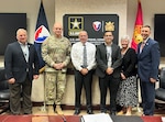 Six people posing for photo with Army sign and two flags behind