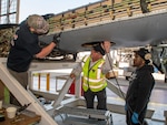 Three men discuss under aircraft wing in industrial space.