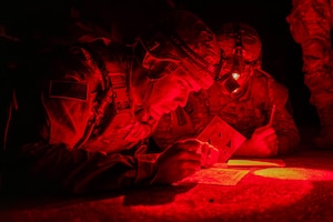 Two soldiers wearing headlamps plot points while lying on the ground in the dark illuminated by red lights.