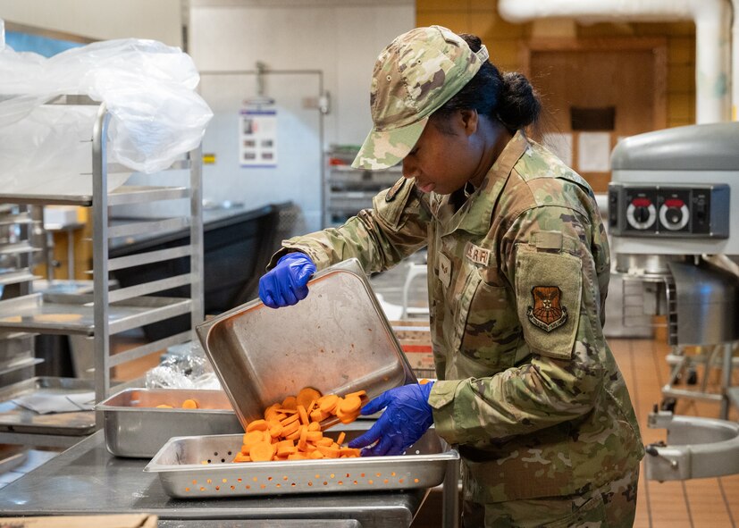 A food services Airman places cooked food into a serving line dish.