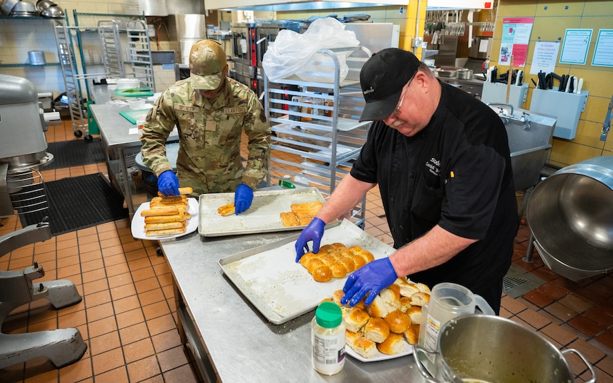 An Airman and a chef in a dining facility kitchen place buttered bread rolls and breadsticks on serving platters.