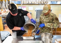 A chef and an Airman in a dining facility kitchen pour soup into a serving tray.