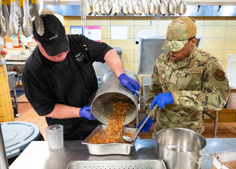 A chef and an Airman in a dining facility kitchen pour soup into a serving tray.