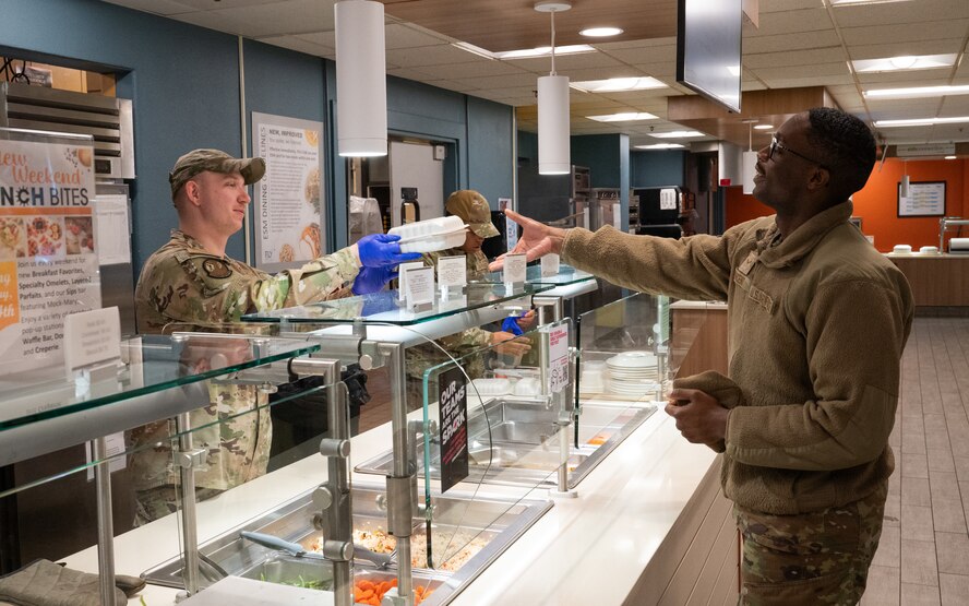 An Airman passes a to-go box of food to another Airman at a dining facility line.