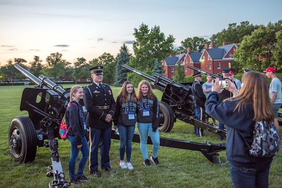 Army soldiers dressed in dark ceremonial uniforms are posing for pictures with audience members next to the Presidential Salute Battery's modified 3-inch M5 anti-tank guns after a Twilight Tattoo show.