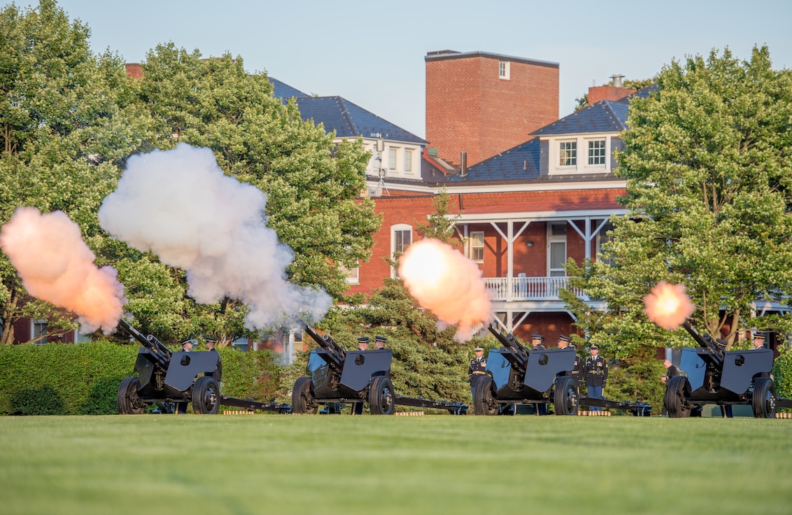 Four black, modified 3-inch M5 anti-tank guns are being fired by Army Soldiers from the Presidential Salute Battery, who are standing near the guns and dressed in dark ceremonial uniforms.