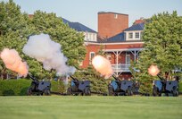 Four black, modified 3-inch M5 anti-tank guns are being fired by Army Soldiers from the Presidential Salute Battery, who are standing near the guns and dressed in dark ceremonial uniforms.