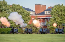 Four black, modified 3-inch M5 anti-tank guns are being fired by Army Soldiers from the Presidential Salute Battery, who are standing near the guns and dressed in dark ceremonial uniforms.