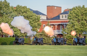 Four black, modified 3-inch M5 anti-tank guns are being fired by Army Soldiers from the Presidential Salute Battery, who are standing near the guns and dressed in dark ceremonial uniforms.