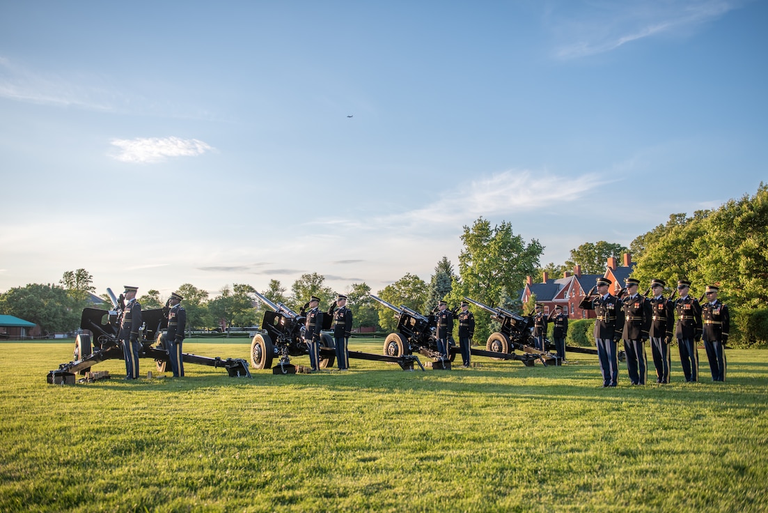 Army Soldiers from the Presidential Salute Battery are dressed in dark blue ceremonial uniforms and standing in formation near four black, modified 3-inch M5 anti-tank guns on a green lawn near a tree line.