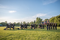 Army Soldiers from the Presidential Salute Battery are dressed in dark blue ceremonial uniforms and standing in formation near four black, modified 3-inch M5 anti-tank guns on a green lawn near a tree line.