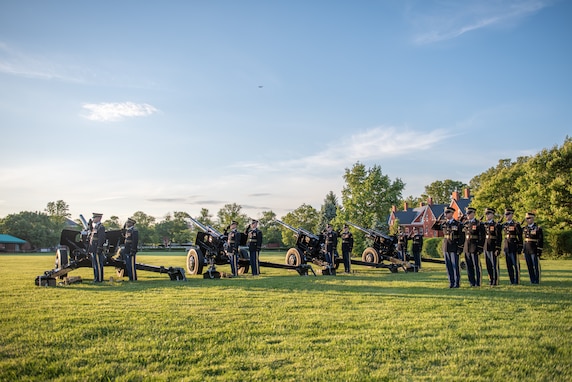 Army Soldiers from the Presidential Salute Battery are dressed in dark blue ceremonial uniforms and standing in formation near four black, modified 3-inch M5 anti-tank guns on a green lawn near a tree line.