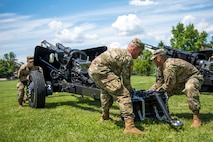 Three Army soldiers with the Presidential Salute Battery wearing combat uniforms are adjusting the position of a black, modified 3-inch M5 anti-tank gun on a green lawn.