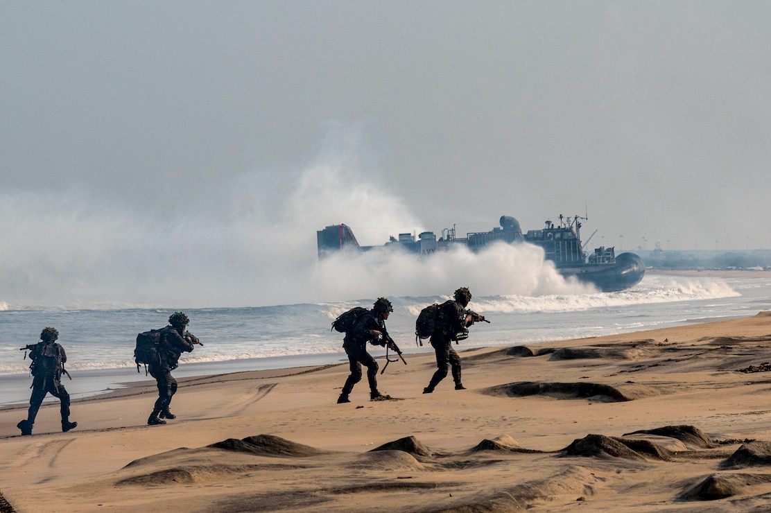 Four Indian solders in tactical gear stand on a beach as a U.S. Navy landing craft splashes ashore in the background.