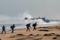 Four Indian solders in tactical gear stand on a beach as a U.S. Navy landing craft splashes ashore in the background.