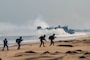 Four Indian solders in tactical gear stand on a beach as a U.S. Navy landing craft splashes ashore in the background.
