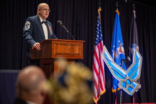 Chief Fitch stands behind a podium wearing formal military uniform beside the U.S., Air Force, and Space Force flags, and a large Air Force insignia