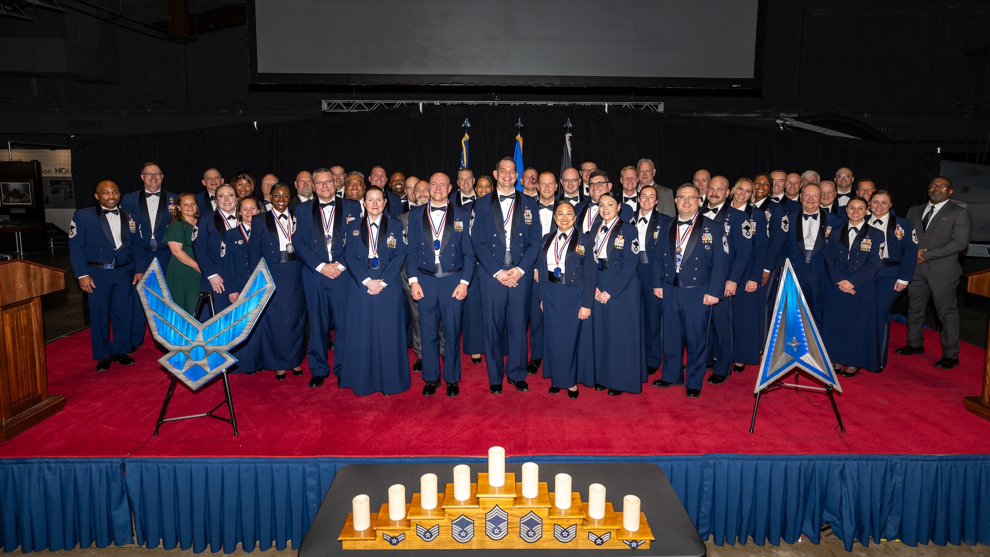 A large group photo of military members in formal wear on stage with large U.S. Air Force and Space Force insignias on either side