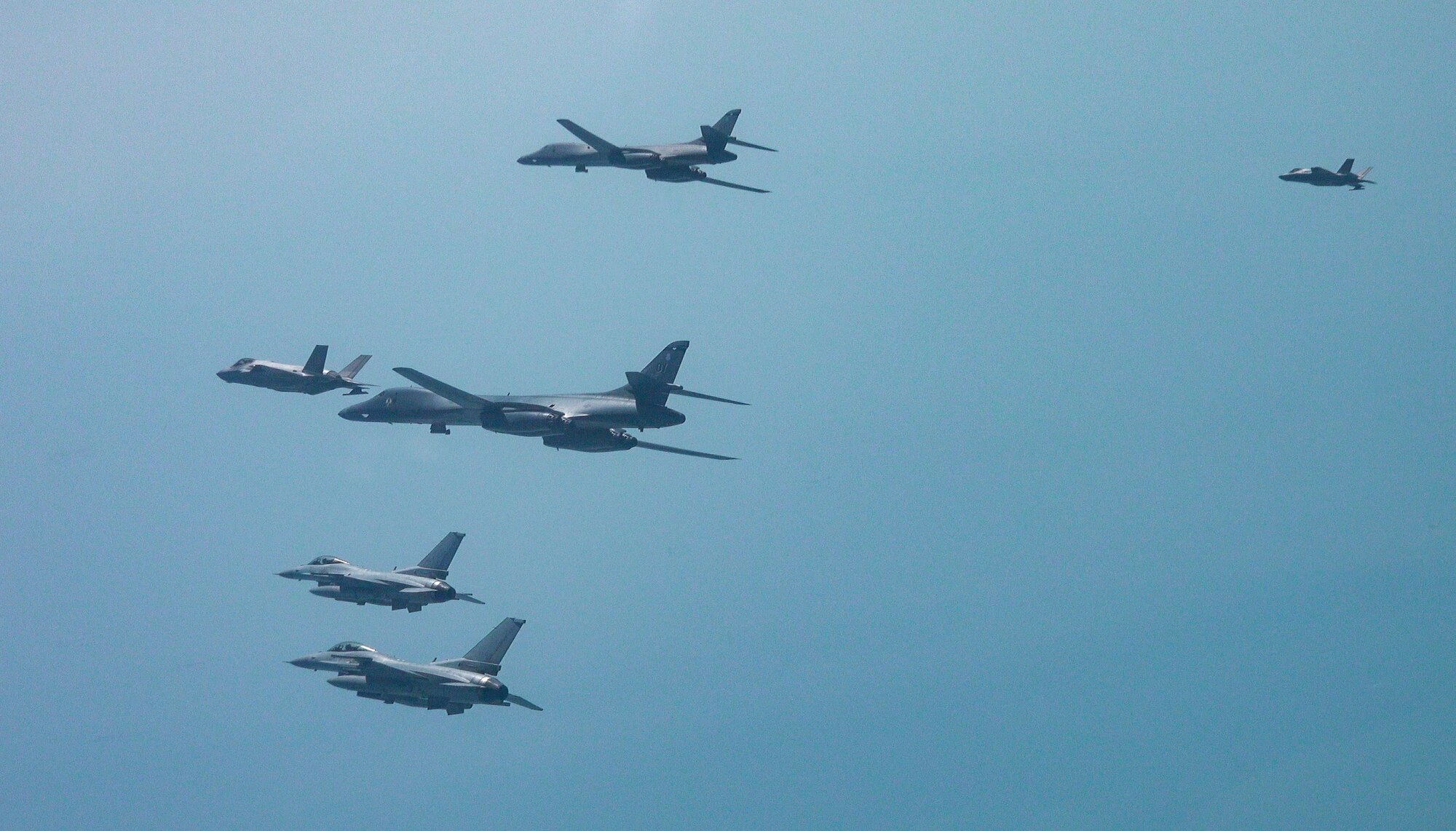 Five aircraft fly in formation from the side view in front of a blue-green hazy sky.