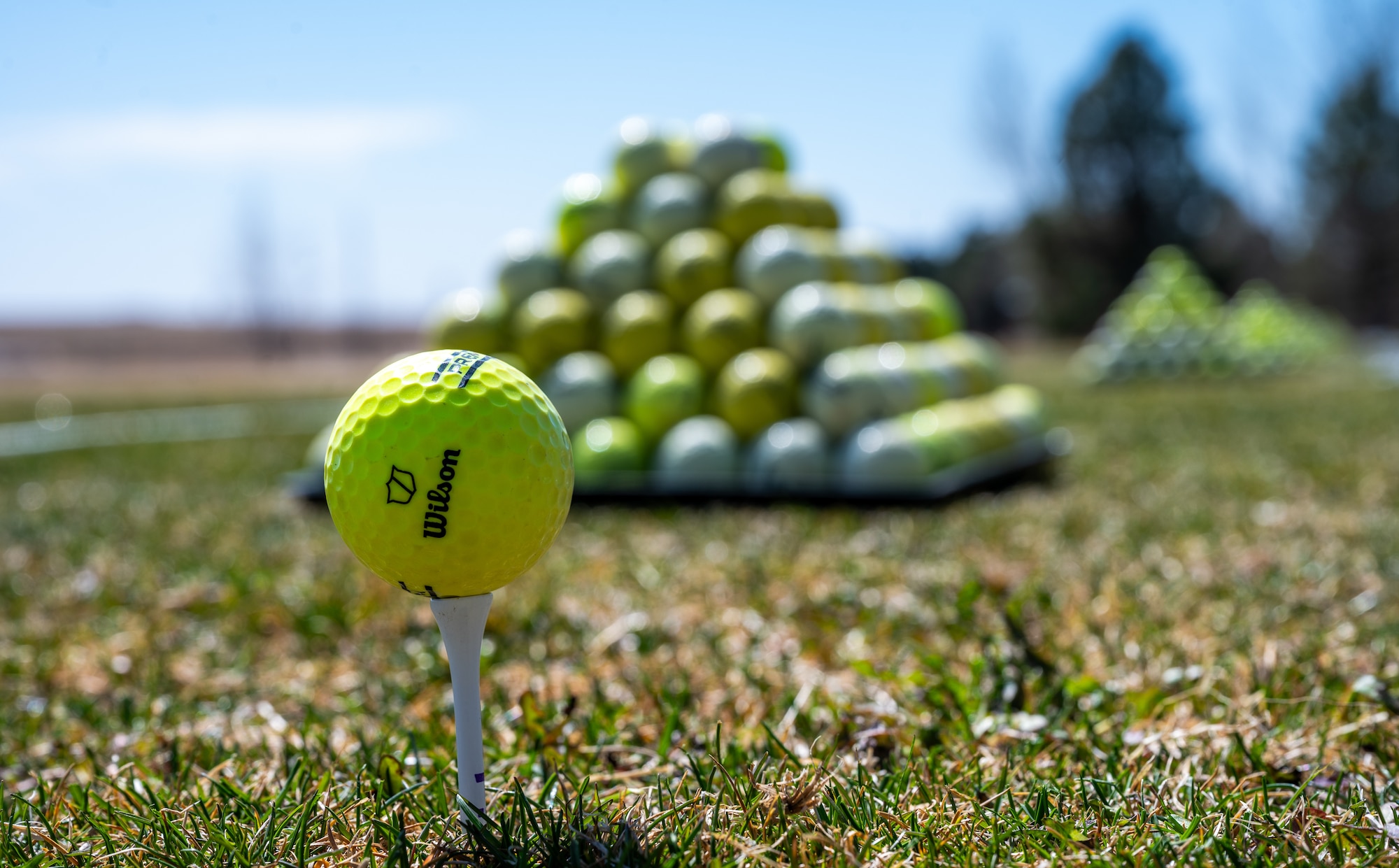 A golf ball rests on a tee over grass with a tower of golf balls stacked behind it.