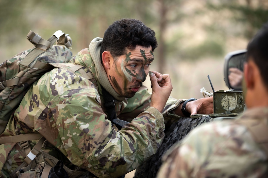 A soldier bends next to a partially visible vehicle while applying face paint with a wooded area in the blurred background.