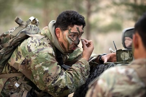 A soldier bends next to a partially visible vehicle while applying face paint with a wooded area in the blurred background.