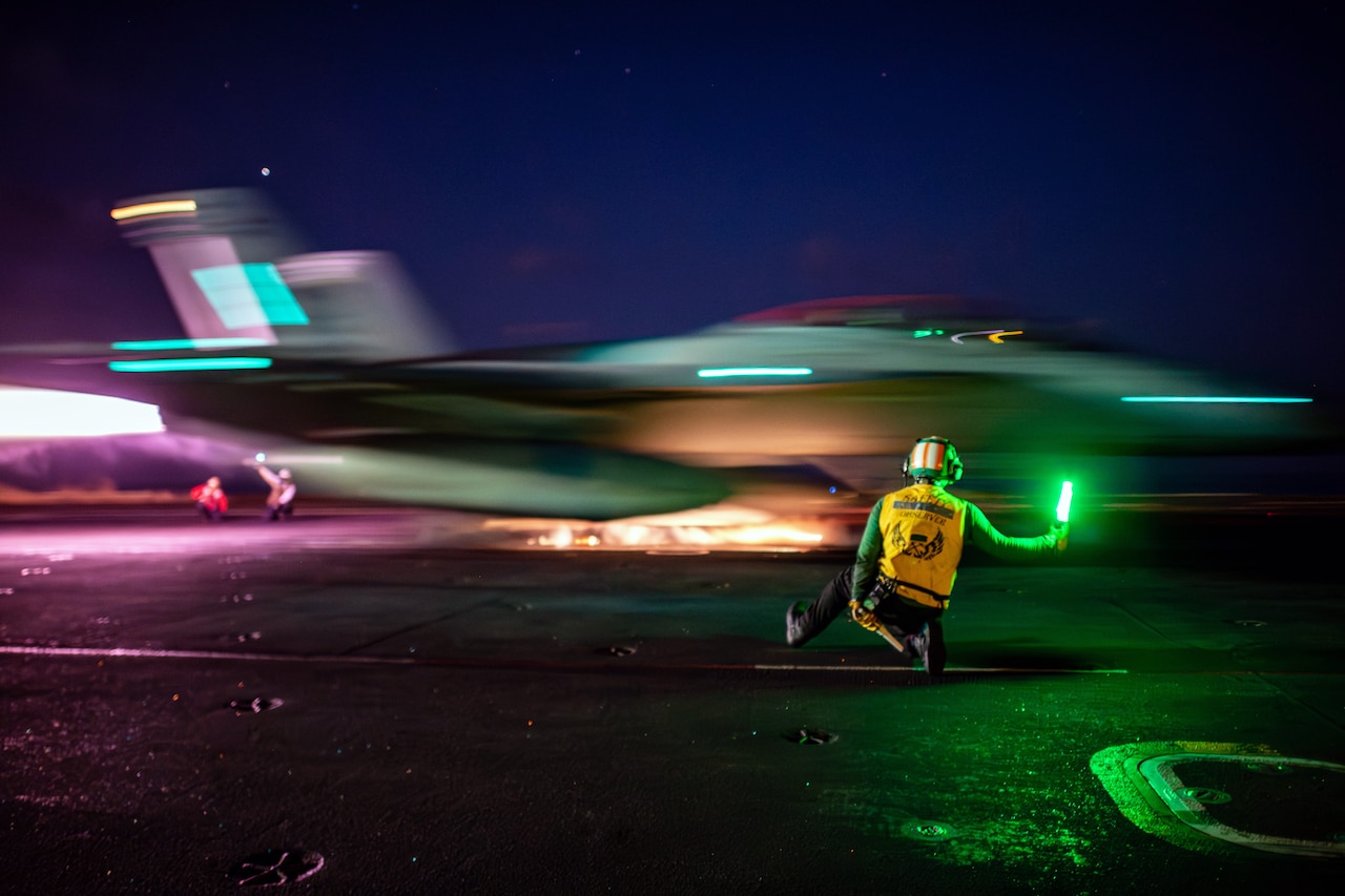A sailor uses a neon baton to signal to a moving aircraft illuminated by bright lights as it prepares to launch from a ship in the dark.