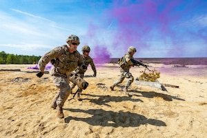 Three soldiers carrying weapons run in sand toward to the right away from clouds of purple smoke on the left.