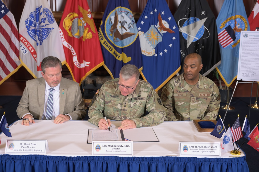 Lt. Gen. Mark Simerly signs a proclamation seated at a table, flanked by Brad Bunn and Chief Master Sgt. Alvin Dyer