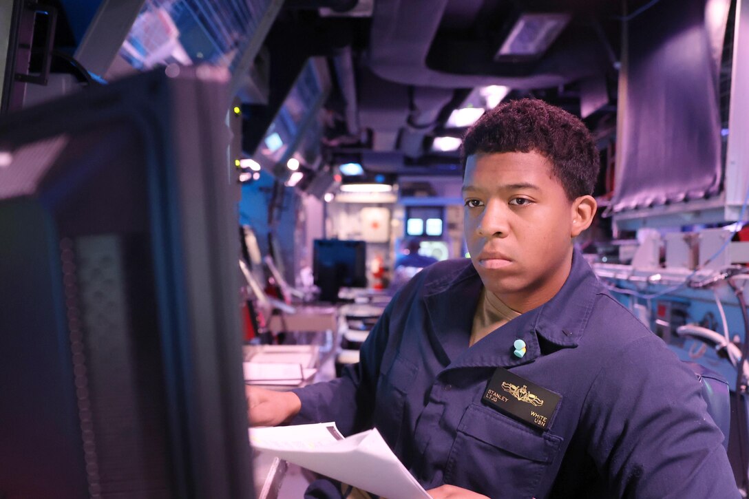 A sailor works on a computer screen in a control room aboard a ship.