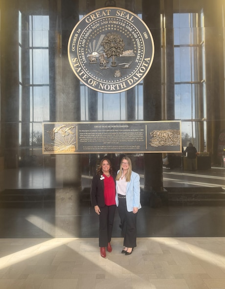 Two women pose for a photo under the Great Seal of North Dakota.