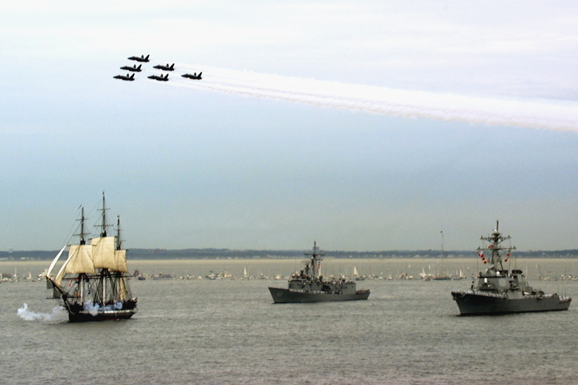 An old ship sails with two modern ships behind and six aircraft flying above.