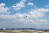 Two aircraft fly in close formation with a white, puffy clouds and a blue sky behind them, over a runway with a town in the distance.